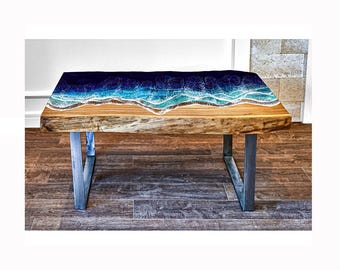 May include: A wooden coffee table with a blue and green epoxy resin top that resembles an ocean wave. The table has a live edge and is supported by two metal legs.