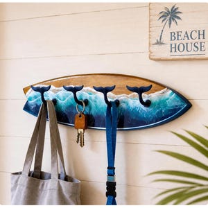 Op de afbeelding: Een wandkapstok in de vorm van een surfplank met een hars golfmotief en vier donkerblauwe walvisstaarthaken. Een klein rechthoekig bordje erboven toont de tekst BEACH HOUSE met een palmboom. Een grijze tas en sleutels hangen aan de haken.