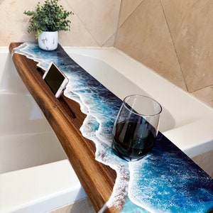 Puede incluir: Una bandeja de baño de madera con un diseño de ola de océano de resina azul y blanca. La bandeja tiene un soporte para teléfono incorporado y un espacio para una copa de vino.