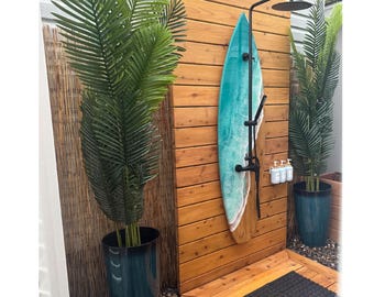 May include: An outdoor shower area featuring a teal surfboard mounted on a wooden wall. The shower has a black showerhead and fixtures, with potted palm plants. The shower area has a wooden floor and a black mat.
