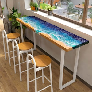 May include: A white bar table with a wooden top featuring a blue and green ocean wave design. Three white bar stools with wooden seats are placed around the table.