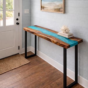 May include: A wooden console table with a turquoise resin river design. The table has a rectangular top and black metal legs. A framed beach scene painting hangs on the wall above the table. The table is 60 inches long.