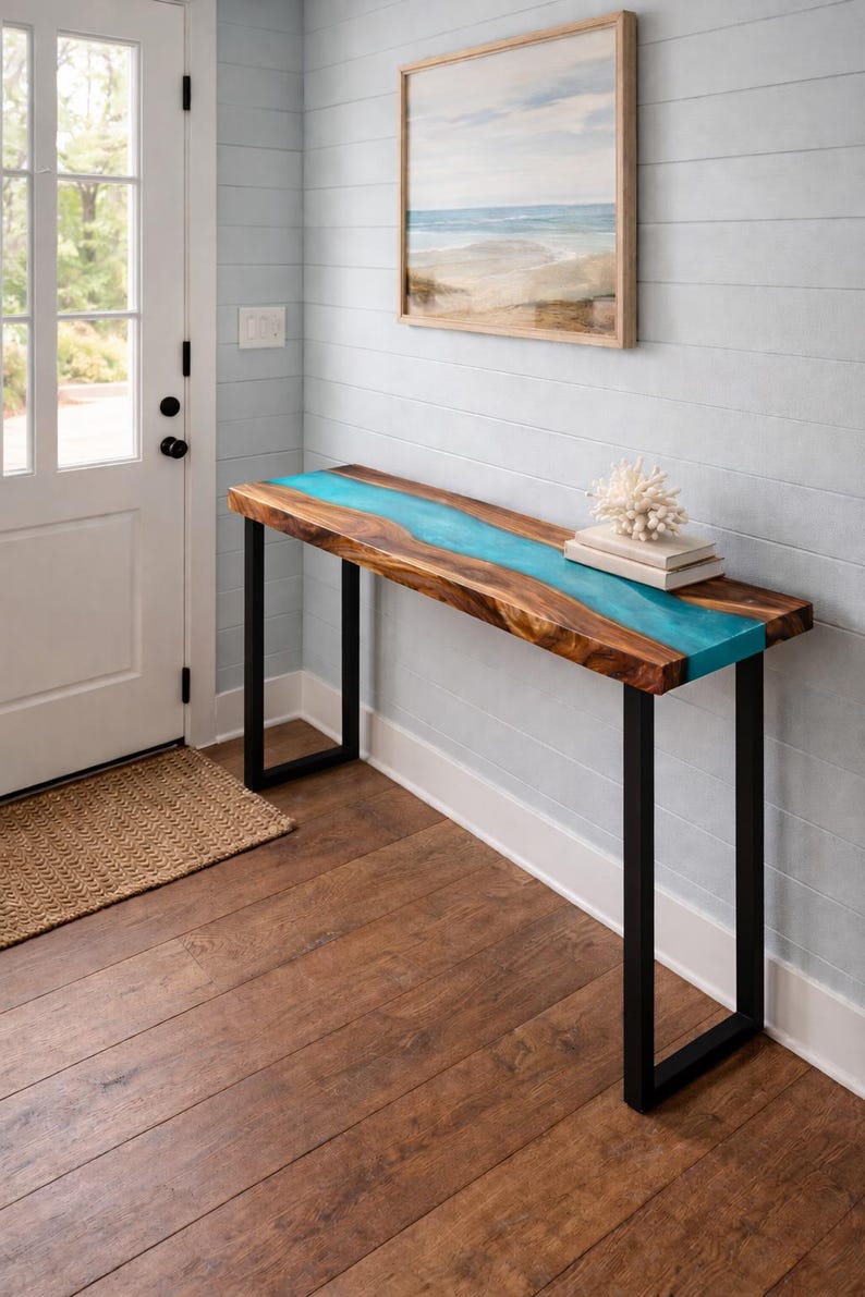 May include: A wooden console table with a turquoise resin river design. The table has a rectangular top and black metal legs. A framed beach scene painting hangs on the wall above the table. The table is 60 inches long.