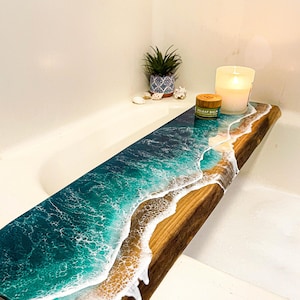 Puede incluir: Un estante de baño de madera con un diseño de ola de océano de resina. El estante está en una bañera blanca y tiene una pequeña planta en maceta, una vela y un tarro de bálsamo encima.