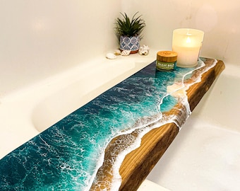 May include: A rectangular wooden bathtub tray featuring a live edge and a resin art design that mimics ocean waves. The surface displays shades of deep blue, turquoise, and white foam, contrasting with the natural brown wood grain texture.