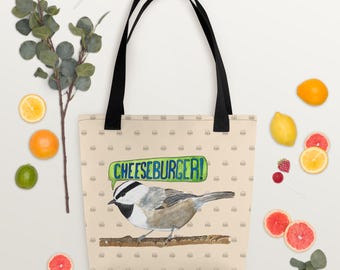 May include: A beige tote bag with black handles featuring a watercolor illustration of a bird perched on a branch. The bag has the text "CHEESEBURGER!" in a green and blue banner, and a pattern of small burger icons.