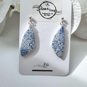May include: A pair of drop earrings featuring a curved, leaf-like shape with a blue and white tile pattern. The earrings are attached to a white display card labelled Diva & Dough Sculpted for the Spotlight and Made by Keli.