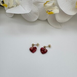 May include: A pair of heart-shaped earrings with a gold post. The earrings are a translucent red with gold flecks. The background is white with white orchid flowers.