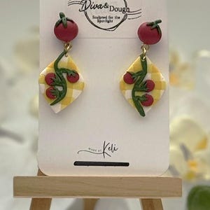 May include: Dangle earrings featuring a red tomato stud and a diamond-shaped charm with a yellow and white checkered pattern, decorated with a green vine and small red tomatoes. The display card reads Diva & Dough Sculpted for the Spotlight and Made by Keli.
