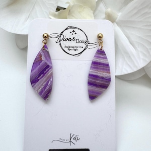 May include: Dangle earrings featuring elongated, diamond-shaped pendants with a purple and white striped marble pattern and gold-tone ball studs. The earrings are attached to a white display card with the text Diva & Dough Sculpted for the Spotlight.