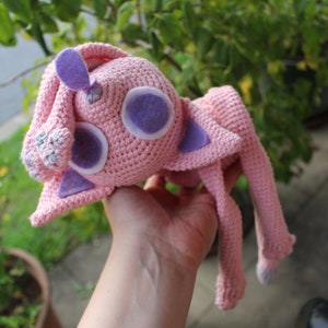 Könnte beinhalten: Ein rosa gehäkeltes Stofftier mit lila Filzohren und -augen. Das Tier hat lange Beine und wird von einer Hand gehalten.