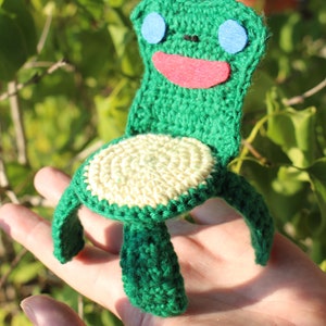 Könnte beinhalten: Ein grüner Häkelstuhl in Form eines Frosches mit rotem Mund und blauen Augen. Der Stuhl hat einen cremefarbenen Sitz.