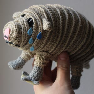 Op de afbeelding: Een gehaakt armadillo knuffeldier met een roze neus en blauwe traanvormige details. Het speelgoed is bruin en beige en heeft een textuur.