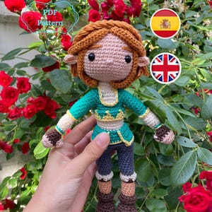 May include: Crocheted amigurumi doll with brown hair, green shirt, and brown boots. The doll is holding its hands out in front of it. The doll is in front of a background of green leaves and red roses.
