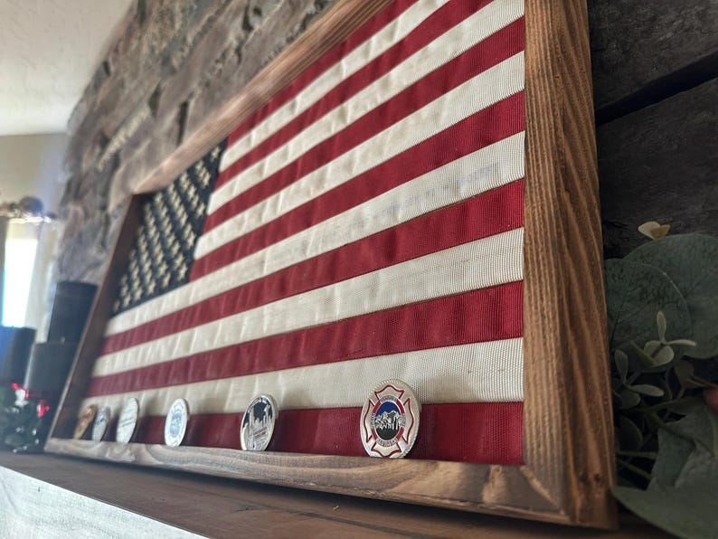 K&ouml;nnte beinhalten: Amerikanische Flagge in einem Holzrahmen. Die Flagge hat rote und wei&szlig;e Streifen und ein blaues Feld mit wei&szlig;en Sternen. Mehrere Gedenkm&uuml;nzen sind darunter ausgestellt und verleihen der patriotischen Dekoration eine pers&ouml;nliche Note.