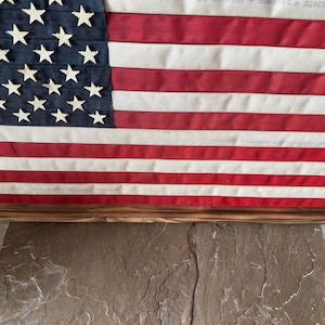 K&ouml;nnte beinhalten: Eine gerahmte amerikanische Flagge mit einem Holzrahmen. Die Flagge zeigt einen blauen Kanton mit wei&szlig;en Sternen und rot-wei&szlig;en Streifen. Die Flagge wird auf einer Steinoberfl&auml;che angezeigt.