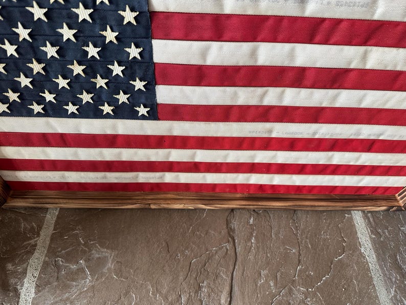 K&ouml;nnte beinhalten: Eine gerahmte amerikanische Flagge mit einem Holzrahmen. Die Flagge zeigt einen blauen Kanton mit wei&szlig;en Sternen und rot-wei&szlig;en Streifen. Die Flagge wird auf einer Steinoberfl&auml;che angezeigt.