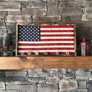K&ouml;nnte beinhalten: Eine gerahmte amerikanische Flagge mit einem Holzrahmen, mit den Sternen und Streifen. Unter der Flagge befinden sich mehrere milit&auml;rische Challenge-M&uuml;nzen. Die Flagge wird auf einem Holzregal vor einer Steinmauer ausgestellt.