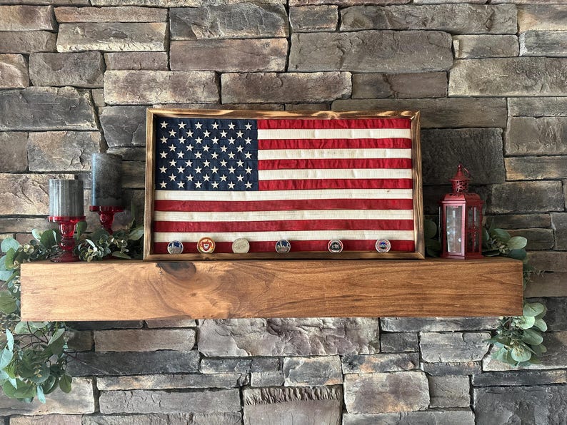 K&ouml;nnte beinhalten: Eine gerahmte amerikanische Flagge mit einem Holzrahmen, mit den Sternen und Streifen. Unter der Flagge befinden sich mehrere milit&auml;rische Challenge-M&uuml;nzen. Die Flagge wird auf einem Holzregal vor einer Steinmauer ausgestellt.