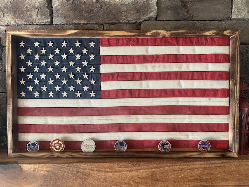 K&ouml;nnte beinhalten: Ein gerahmter amerikanischer Flagge mit einem Holzrahmen. Die Flagge hat rote und wei&szlig;e Streifen und ein blaues Feld mit wei&szlig;en Sternen. Darunter befinden sich sechs runde M&uuml;nzen mit verschiedenen Designs.