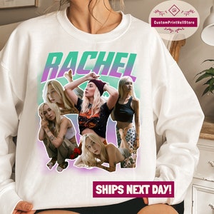 May include: White sweatshirt with a green and purple gradient background. The text "RACHEL" is printed in large, bold letters. The sweatshirt features a collage of four women with different hairstyles and clothing.