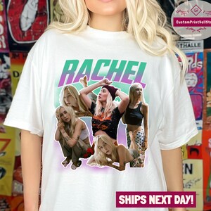 May include: White t-shirt featuring a graphic print of the name "Rachel" in a green and purple gradient. The graphic also includes four women with various hairstyles and outfits.