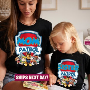 May include: Two black t-shirts with a Paw Patrol theme. The larger shirt says "Mom Patrol" with a Paw Patrol logo and the smaller shirt says "Sister Patrol" with a Paw Patrol logo.
