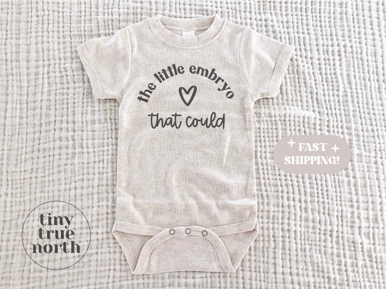 May include: A light beige baby onesie with short sleeves and a snap closure. The text "the little embryo that could" is printed in black, with a heart symbol. The brand logo "tiny true north" is also visible.