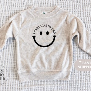 May include: A light grey sweatshirt with the text "I fart like my dad" and a black smiley face graphic.