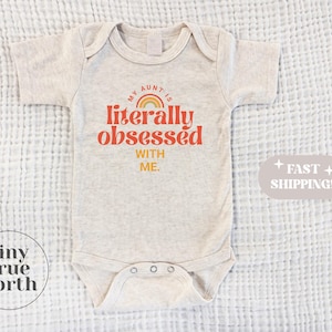 Könnte beinhalten: Ein hellgrauer Baby-Body mit dem Text "MY AUNT IS literally obsessed WITH ME." in Orange und Gelb. Über dem Wort "literally" befindet sich ein Regenbogen. Der Body hat kurze Ärmel und Druckknöpfe.