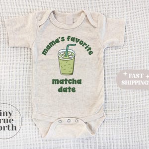 May include: A light beige baby onesie with the text "mama's favorite matcha date" in green. The design includes a green matcha drink illustration with a straw. The onesie has short sleeves and three snaps at the bottom.