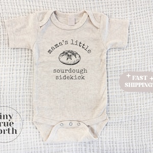 May include: A beige baby onesie with a black graphic of a sourdough bread loaf and the text "mama's little sourdough sidekick".