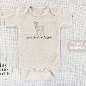 May include: A beige baby onesie with a black and white illustration of a goat and the text "new kid in town".