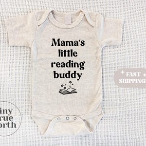 Pode incluir: Um body de bebé bege claro com o texto "Mama's little reading buddy" em preto. Um gráfico de um livro aberto com estrelas está abaixo do texto. O body tem mangas curtas e fechos de pressão. As palavras "Fast Shipping!" estão num oval cinza claro.