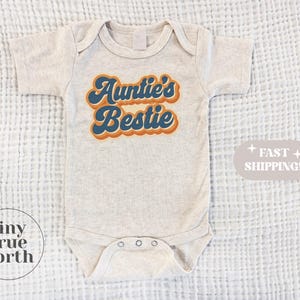 Puede incluir: Un body de bebé beige claro con el texto "Auntie's Bestie" en una fuente retro con acentos azules y naranjas. El body tiene mangas cortas y corchetes en la parte inferior. La imagen está sobre un fondo blanco texturizado.