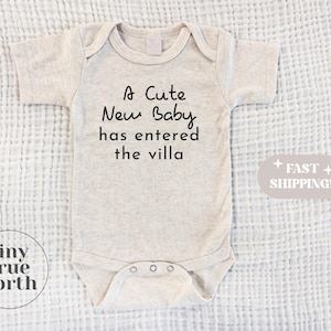 May include: A beige baby onesie with the text "A Cute New Baby has entered the villa" printed in black.