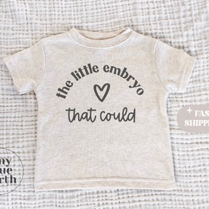 May include: A light beige toddler t-shirt with the text "the little embryo that could" in black, with a heart symbol. The shirt is short-sleeved and made of a soft material.