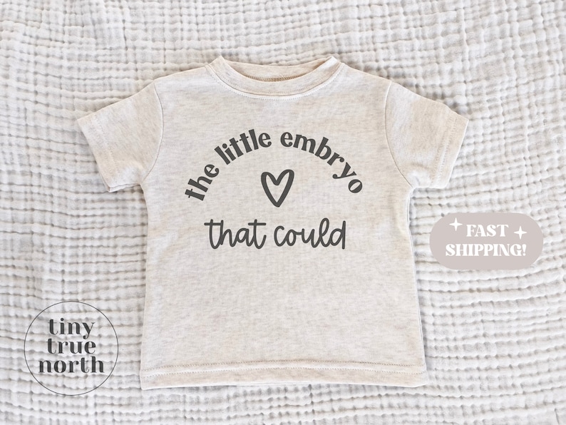 May include: A light beige toddler t-shirt with the text "the little embryo that could" in black, with a heart symbol. The shirt is short-sleeved and made of a soft material.