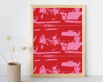 May include: A framed pop art print featuring a repeating pattern of a woman in a car. The artwork uses a bold color palette of red and pink, with a light wood frame. The print is displayed on a white surface.