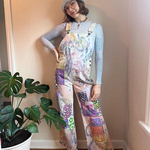 Op de afbeelding: Een persoon draagt een lichtblauwe gebreide muts, een lichtblauw shirt met lange mouwen en een overall van patchwork met een tie-dye patroon. De overall heeft verschillende kleurrijke patches die erop zijn genaaid. De persoon staat voor een plant in een pot.