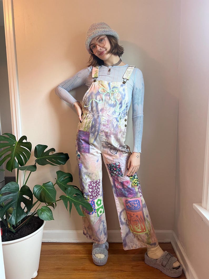 Op de afbeelding: Een persoon draagt een lichtblauwe gebreide muts, een lichtblauw shirt met lange mouwen en een overall van patchwork met een tie-dye patroon. De overall heeft verschillende kleurrijke patches die erop zijn genaaid. De persoon staat voor een plant in een pot.