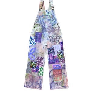 Op de afbeelding: Een lichtblauwe spijkerbroek met bretels met een tie-dye patroon en kleurrijke patchwork details. De spijkerbroek heeft een vlinderontwerp en bloemenpatronen.