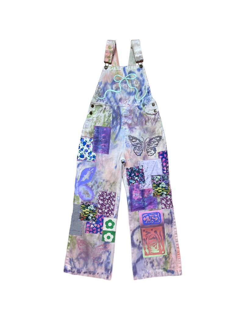 Op de afbeelding: Een lichtblauwe spijkerbroek met bretels met een tie-dye patroon en kleurrijke patchwork details. De spijkerbroek heeft een vlinderontwerp en bloemenpatronen.