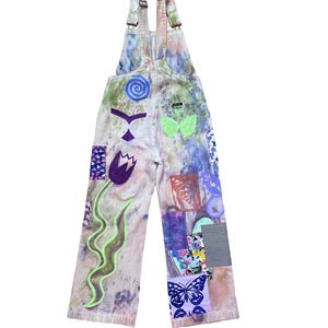Op de afbeelding: Een lichtblauwe spijkerbroek met bretels met een tie-dye patroon en verschillende kleurrijke patches, waaronder een bloem, een vlinder en een swirl-ontwerp.