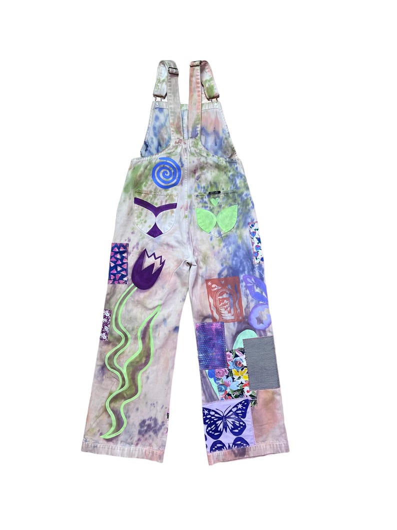 Op de afbeelding: Een lichtblauwe spijkerbroek met bretels met een tie-dye patroon en verschillende kleurrijke patches, waaronder een bloem, een vlinder en een swirl-ontwerp.