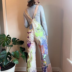 Op de afbeelding: Een persoon draagt een lichtblauw lang mouwen shirt en een paar witte overalls met een kleurrijk patchwork ontwerp. De overalls hebben een verscheidenheid aan patronen, waaronder bloemen, vlinders en abstracte vormen. De persoon draagt een lichtblauwe beanie en een bril.