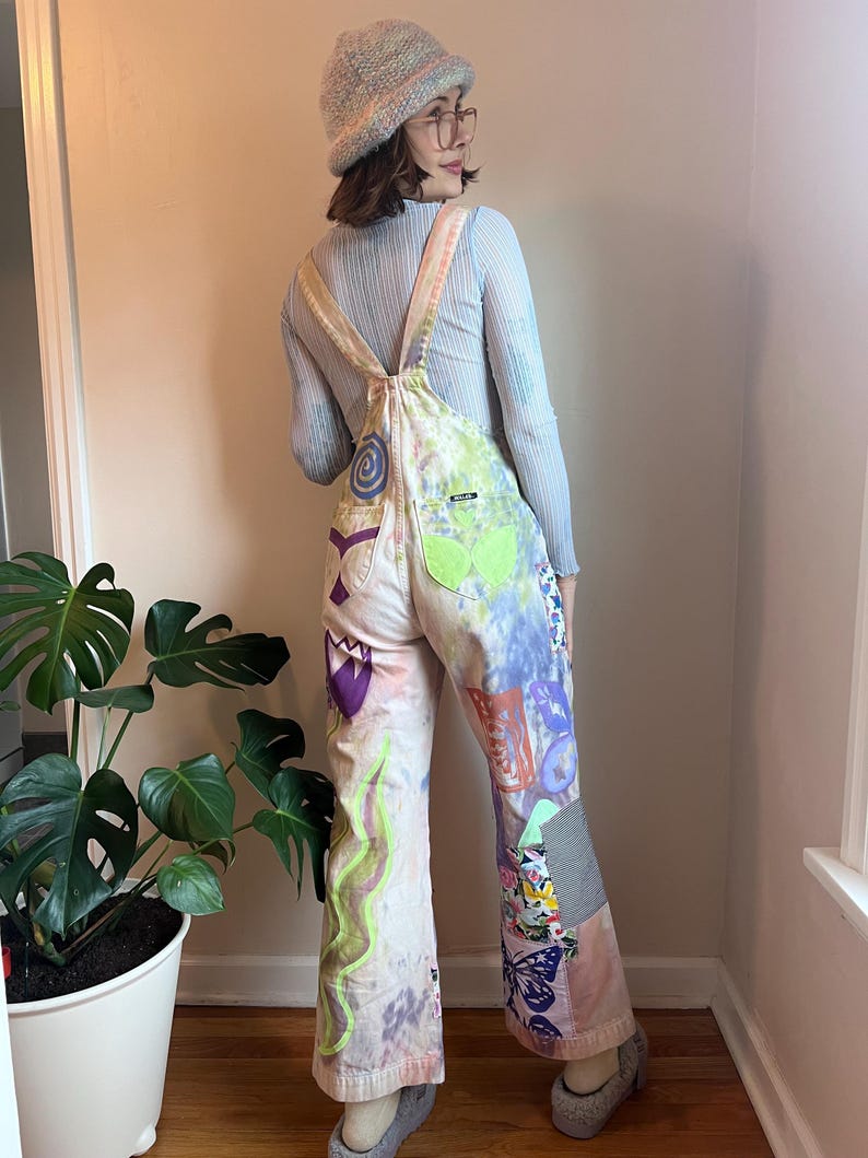 Op de afbeelding: Een persoon draagt een lichtblauw lang mouwen shirt en een paar witte overalls met een kleurrijk patchwork ontwerp. De overalls hebben een verscheidenheid aan patronen, waaronder bloemen, vlinders en abstracte vormen. De persoon draagt een lichtblauwe beanie en een bril.