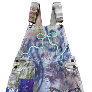 Op de afbeelding: Tie-dye denim overall met een lichtblauwe geborduurde strik en een vlinderpatch. De overall heeft een patchwork-ontwerp met bloemen- en geometrische patronen.