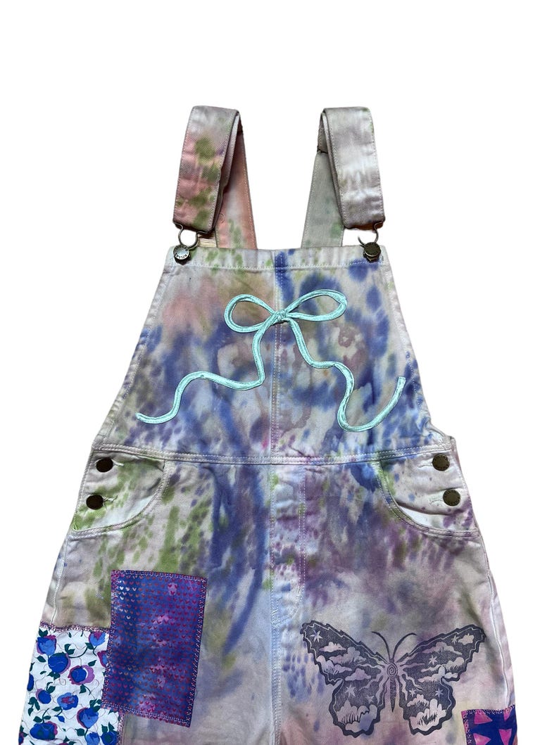 Op de afbeelding: Tie-dye denim overall met een lichtblauwe geborduurde strik en een vlinderpatch. De overall heeft een patchwork-ontwerp met bloemen- en geometrische patronen.