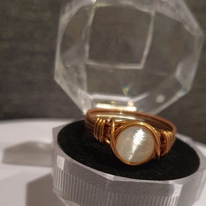 May include: A handcrafted ring with a copper-coloured wire band and a round, white stone. The ring is displayed inside a clear, faceted, plastic box with a black interior. The ring's design features wrapped wire detailing.